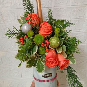 Centro de mesa navideño con rosas naranjas, follaje verde y detalles de esferas festivas, decorado con una vela dorada.