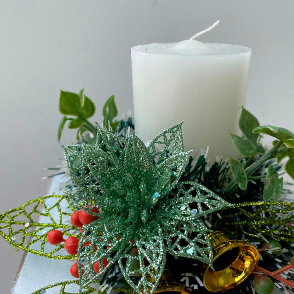 Centro de mesa navideño con vela blanca, hojas verdes y campanas doradas.